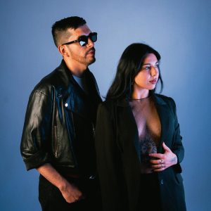 Aparece un fondo tenue color celeste con dos personas mirando hacia la derecha: un hombre con barba, vestido con lentes oscuros, chaqueta de cuero negra y pantalones negros con su mano en el bolsillo. A la derecha una mujer con pelo negro y suelto con un blazer negro y un pantalón negro y su mano en su vientre