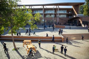 La imagen muestra un día soleado en el patio del colegio Miguel Cruchaga Tocornal. Con estudiantes en distintas ubicaciones. Las salas al fondo, un árbol a la izquierda y unas mesas de madera en el mismo sector