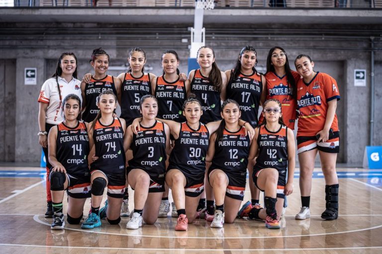 La imagen está ubicada en una cancha de básquetbol. Aparecen 13 jugadoras, 7 paradas y 6 en arrodilladas para una foto grupal, con uniformes de negro y naranjo. A la izquierda acompañando a las jugadoras está su entrenadora con polera blanca y pantalones negros.
