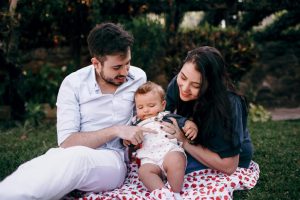 En un parque están sentados en una manta un hombre, vestido con camisa y pantalones blanco, en el centro hay un bebe con un body blanco y a la derecha hay una mujer con una polera verde y pelo suelto. Ambos tienen en sus brazos al bebé.